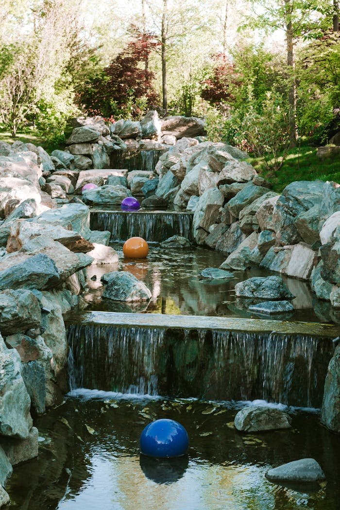 Garden stream with cascading waterfalls and colorful balls surrounded by lush flora.